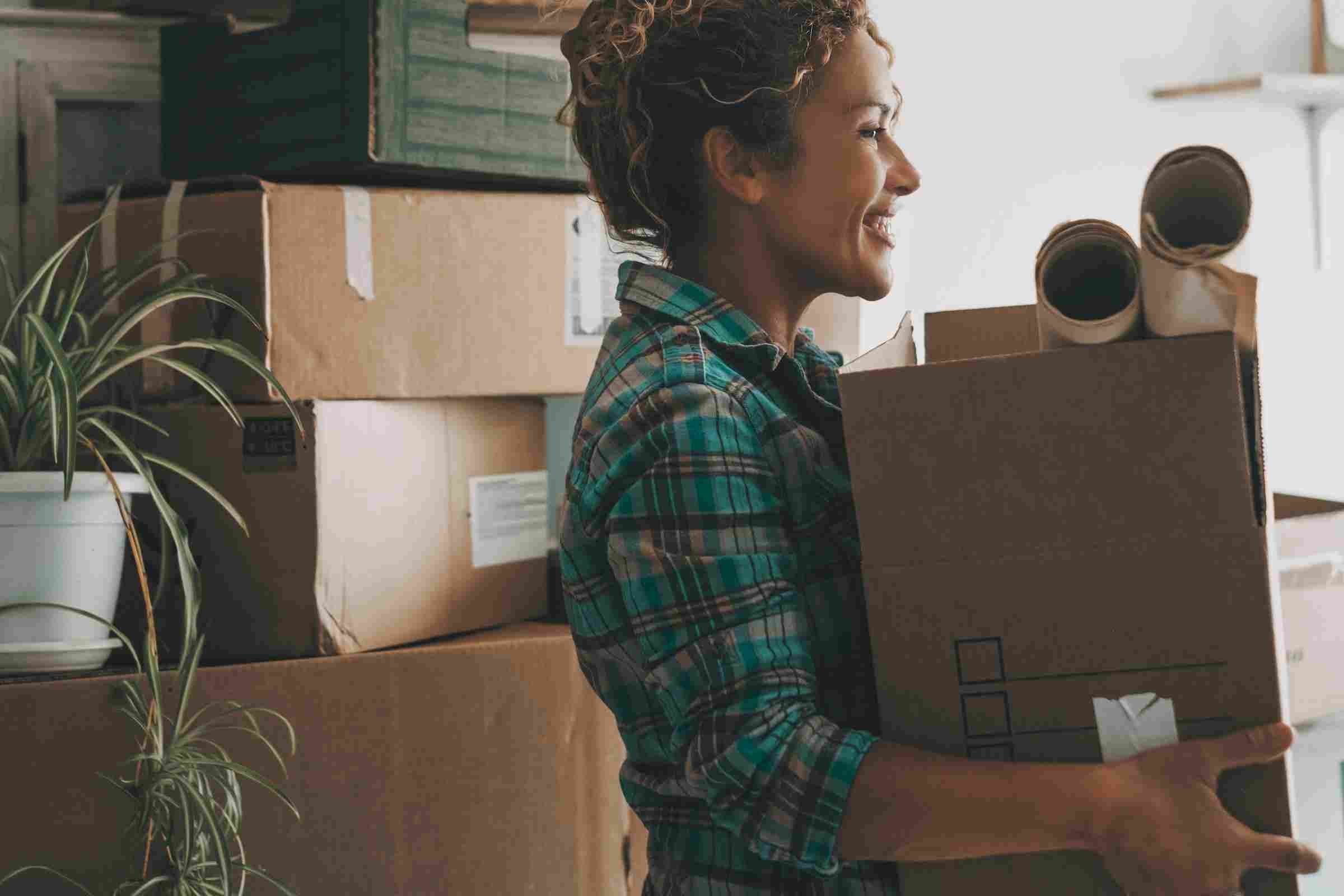 Smiling woman holding a box and packing supplies, surrounded by moving boxes and a potted plant.