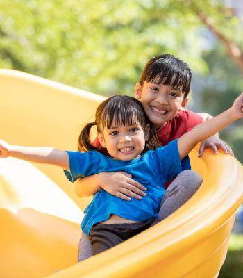 Two smiling children sliding down a yellow slide at a playground, arms outstretched, enjoying themselves.
