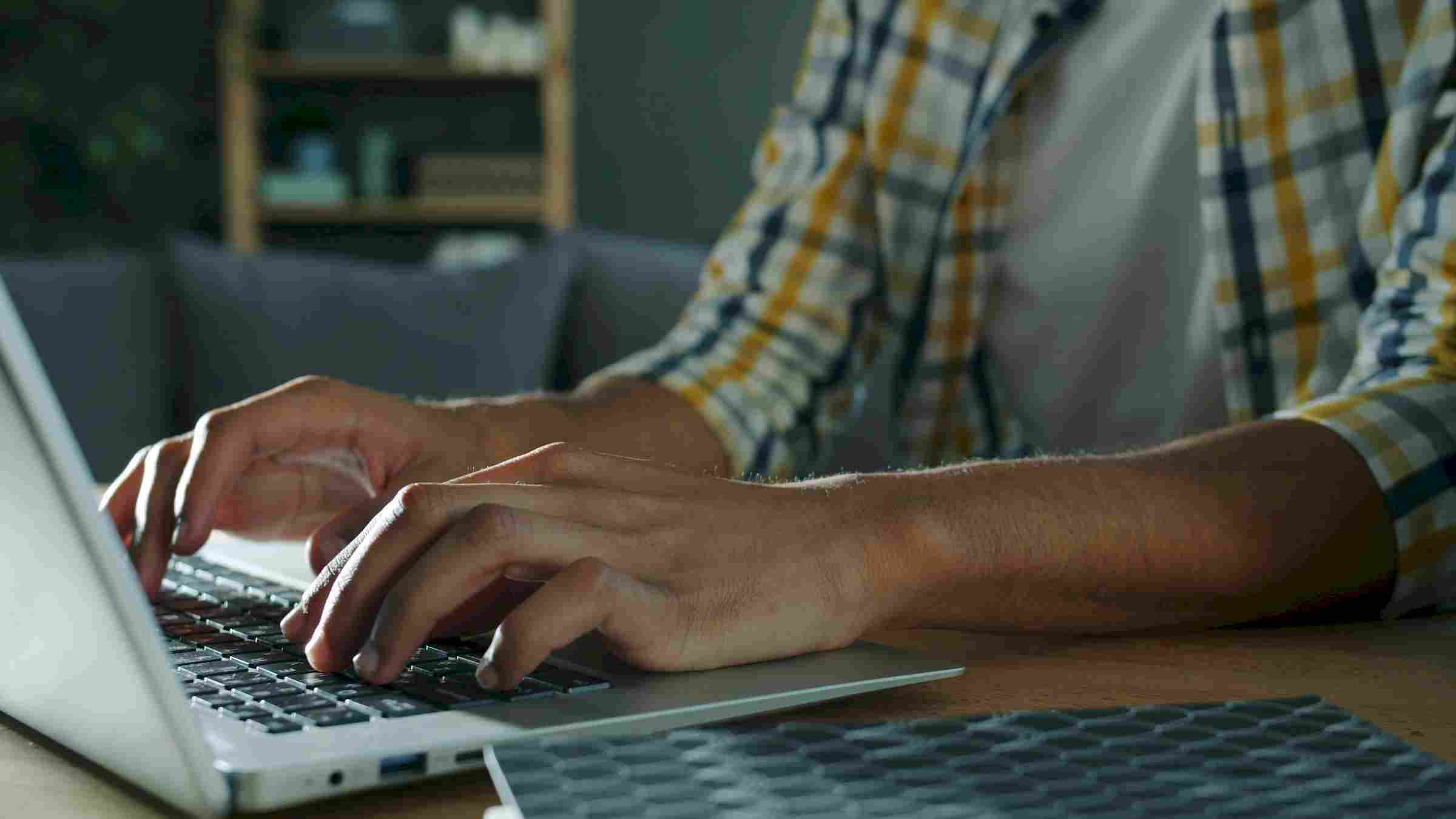 Person typing on a laptop at a desk, wearing a plaid shirt, with a keyboard visible in the foreground.